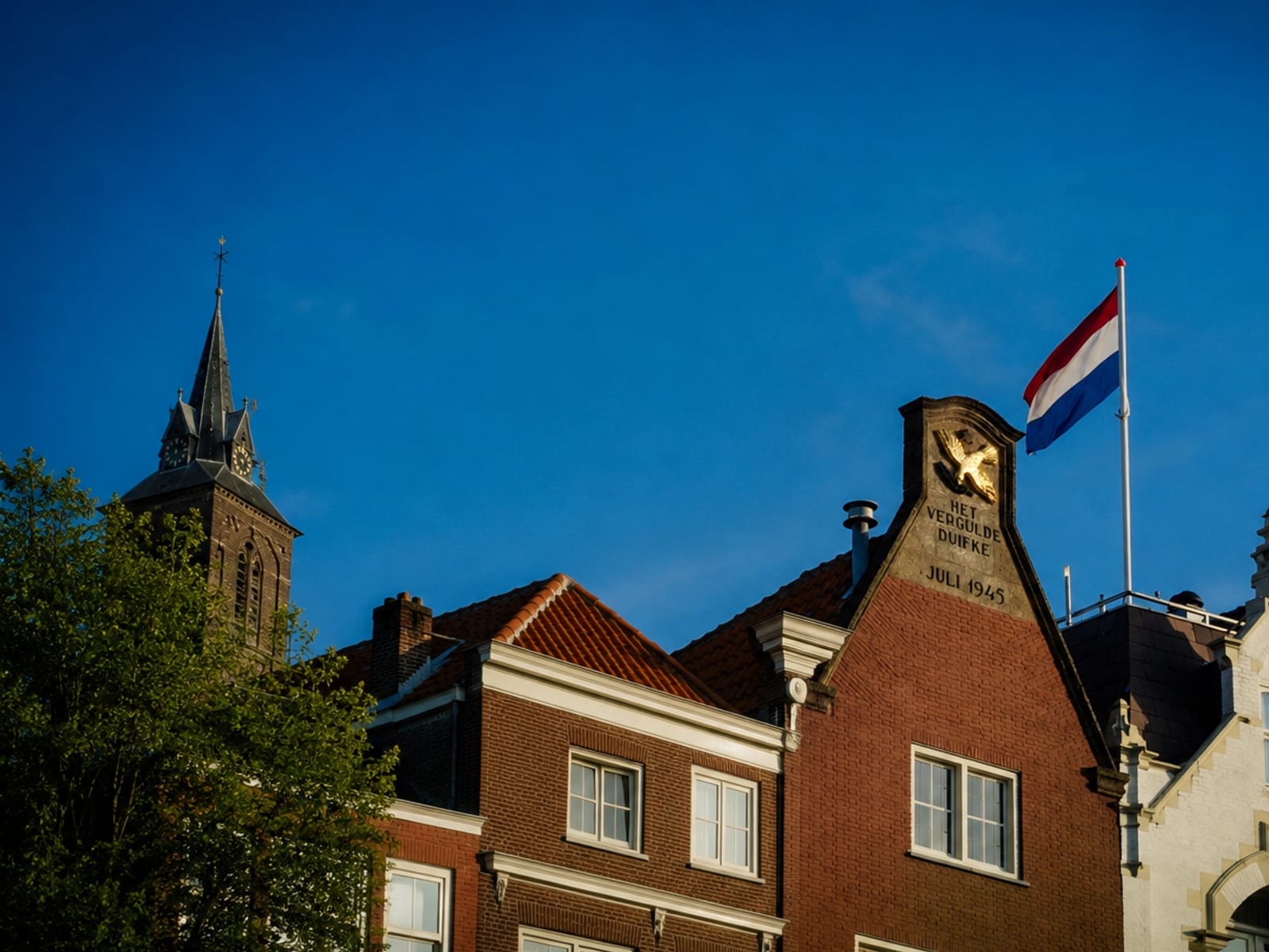 Nederlandse trapgevels met het opschrift 'Het Vergulde Duifke, juli 1945', een kerktoren en de Nederlandse vlag